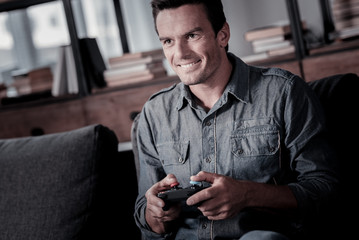 Favorite leisure activity. Handsome adult guy wearing casual attire relaxing on a sofa and smiling cheerfully while playing video games at home.