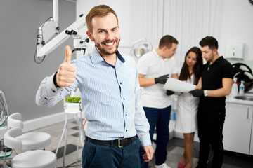 Young attractive male client of a dentistry is smiling to the camera and showing thumbs up being...