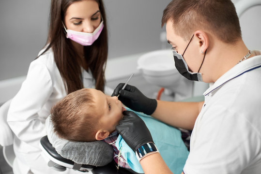 A child is having his teeth attended by a male dentist in a black mask and gloves being assisted by a pretty female in a white modern dental clinic.