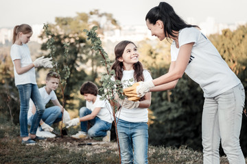 Doing good things. Joyful cute nice girl holing a young tree and smiling while being happy about...