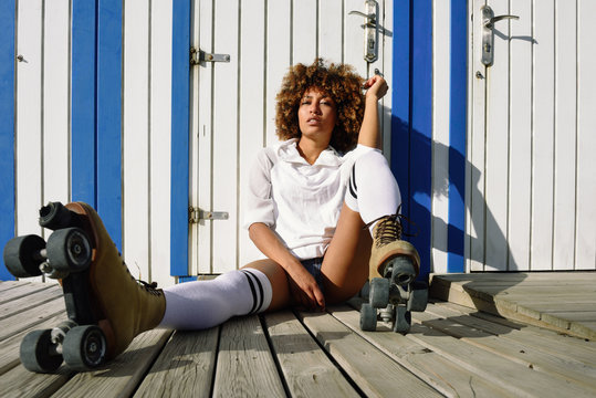 Young black woman on roller skates sitting near a beach hut.