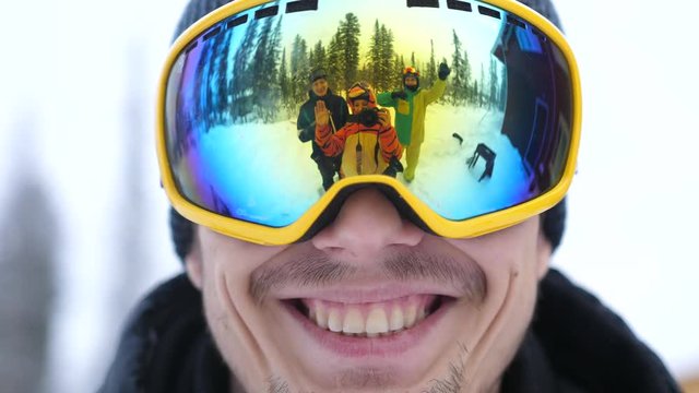 Portrait of smiling man in skiing glasses in deep beautiful forest and cheerful friends waving their hands and reflected in his glasses during snowfall. slow motion. 3840x2160