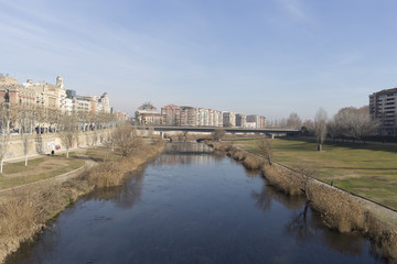 Obraz premium Blue river plain on a green nature park on Lleida city, with architectural details on a the skyline and solid blue sunny sky background