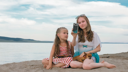 Two teenage are sitting on a sandy beach. They wear sunglasses. Sisters talk to each other and selfie on phone. They have two dogs. Girls correspond, playing, on the Internet.