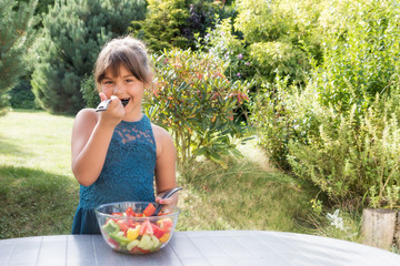 Smiling little girl is tasting salad outdoors. The girl is looking at the camera.