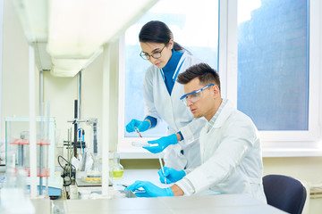 Fototapeta premium Team of ambitious researchers wearing white coats and rubber gloves carrying out experiment while gathered together at modern laboratory