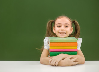 Happy schoolgirl with pile books near empty green chalkboard. Space for text