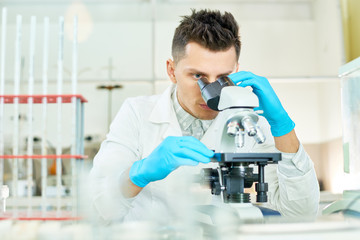 Portrait shot of concentrated young researcher wearing white coat and rubber gloves looking through microscope while working on ambitious project, interior of modern lab on background