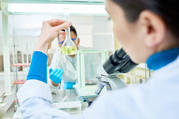 Over shoulder view of young dark-haired chemist holding flask with yellow solution in hand while sitting at desk, interior of modern lab on background