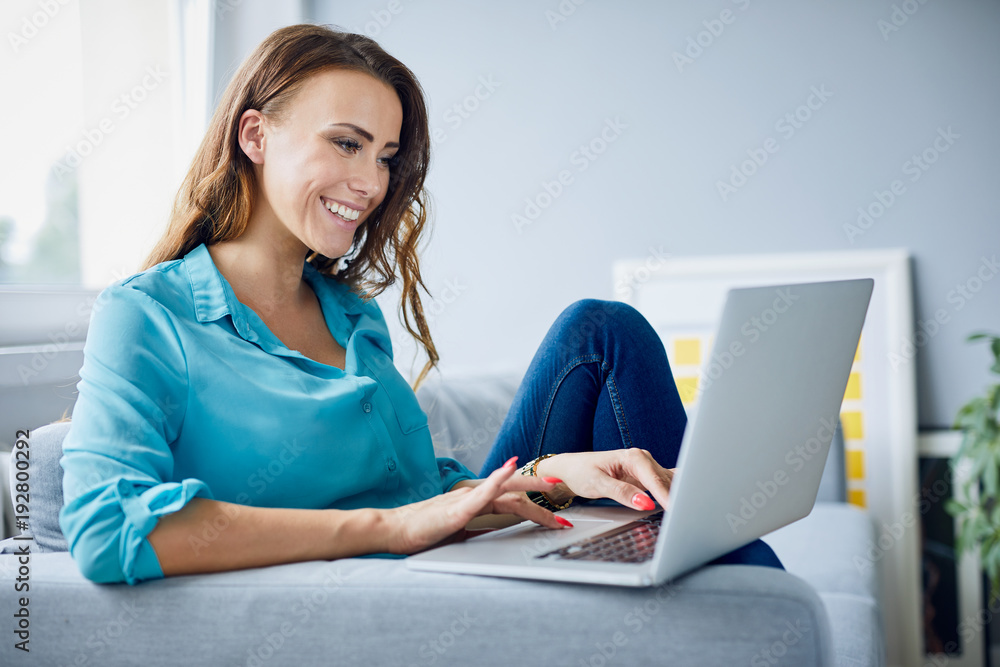 Wall mural side view of a beautiful young woman using laptop and sitting on sofa at home