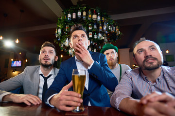 Group of concentrated friends watching football match while sitting at bar counter and drinking beer, bearded man wearing leprechaun costume standing behind them