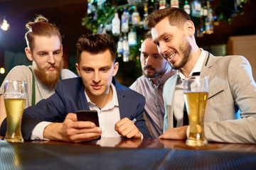 Group of bearded colleagues gathered together in pub after hard working day, drinking beer and watching video on smartphone