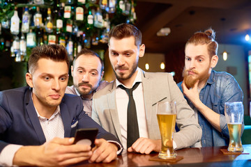 Group of surprised bearded men gathered together in bar, drinking lager and reading news with help of smartphone