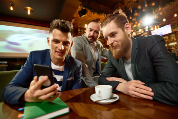 Group of friends watching funny video on smartphone and enjoying fragrant coffee while gathered together in modern cafe