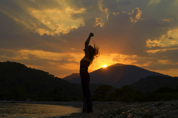 Girl stands on the sea beach with hands up. Hair falls. Sunset at the seaside with mountains on background. Sun breaks through the clouds. A stream of light in evening sky. Woman silhouette. Summer.
