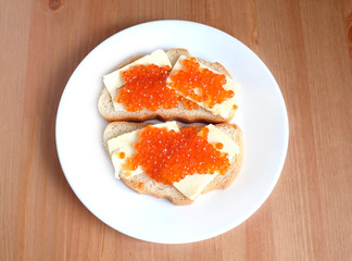 Two sandwiches with butter and red salmon caviar on white bread lies on white round plate on wooden background top view