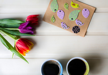Tulips on wooden background with two cups of coffee. Invitation postcard for mother's day or international women's day. Spring paper birds on craft paper envelope. Handmade origami. Punchy pastels