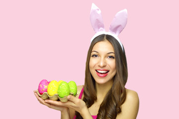 Obraz na płótnie Canvas Easter concept. Beautiful young woman with bunny ears holdings egg carton of colorful Easter eggs isolated on pink background. Copy space.