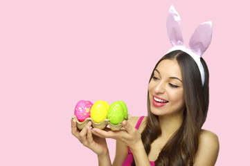 Easter woman concept. Cute girl with bunny ears looking to her egg carton of colorful Easter eggs isolated on pink background. Copy space.