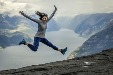 Portrait of young and sporty woman jumping on the Preikestolen plateau. Scenic fjord on background, Norway.