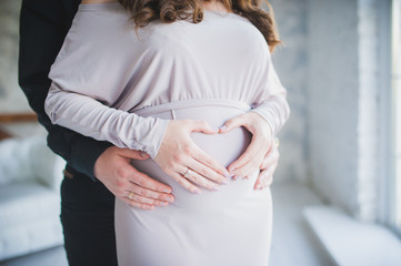 A pregnant girl hugs her tummy. A man is hugging the belly of a pregnant woman. Pregnant woman in a dress. Hands in the form of a heart on the stomach. Walking on the tummy of a pregnant woman