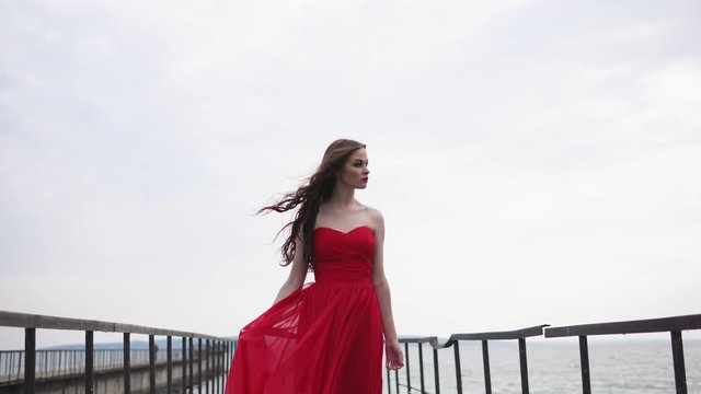 Alone woman is wearing red chiffon dress is looking dramatically on sea. She is standing in a middle of a long pier in windy weather, camera is approaching to her