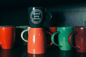 A dark indigo coffee cup named Time For Coffee standing on an orange cup with colorful background of a group of mugs, on a black shelf