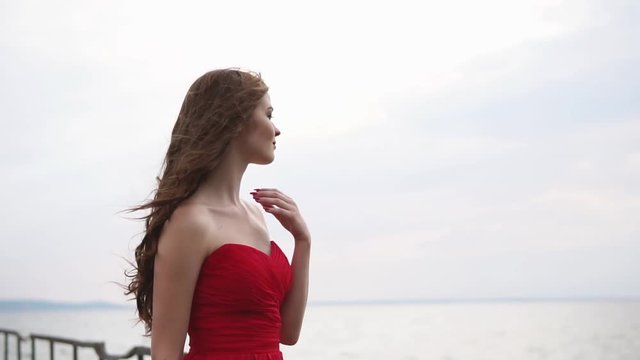 Thoughtful beautiful woman is looking on sea horizon in cloudy weather. Wind is moving her hair and chiffon dress, girl is touching her hair