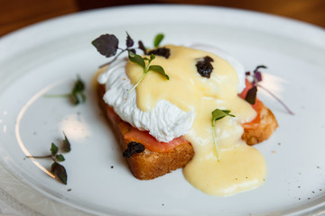 A horizontal image of eggs benedict on a white plate. A grilled toast with salmon, poached egg, hollandaise sauce, truffles and greenery. Selective focus.