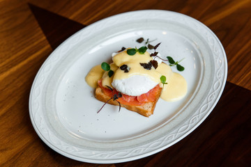 A horizontal image of eggs benedict on a white plate. A grilled toast with salmon, poached egg, hollandaise sauce, truffles and greenery. Selective focus.