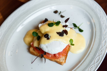 A horizontal image of eggs benedict on a white plate. A grilled toast with salmon, poached egg, hollandaise sauce, truffles and greenery. Selective focus.