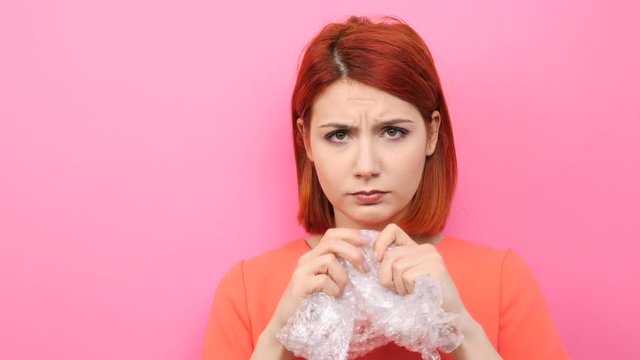 Redhead woman pops a bubble wrap on pink background to calm herself