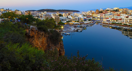 Naklejka premium Beautiful southern town of Agios Nikolaos at summer evening. Boats swing on the water of the lake Voulismeni at the pier with evening lights. Crete Island. Greece