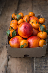 A cardboard box of fresh winter fruits with red oranges and mini tangerines. On a wooden background