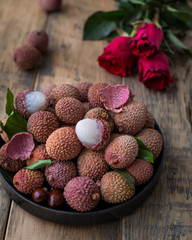 Still life of fresh lychee fruits in a black plate on a wooden background. Rustic style. Decorated with red roses