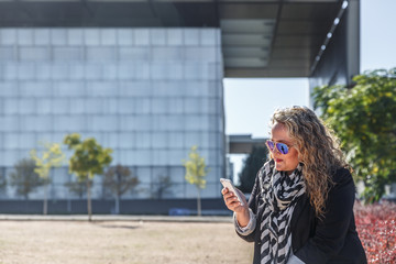 A mature blond woman looks attentively at her mobile phone in the middle of a park