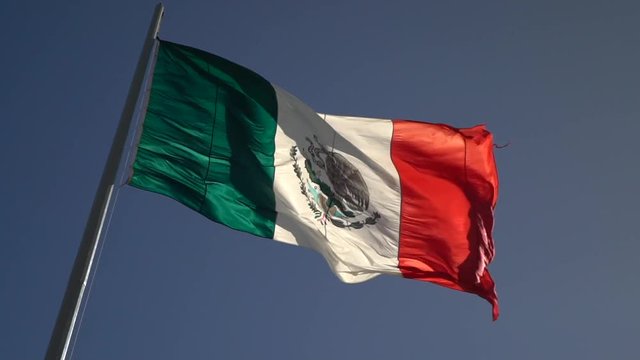 Facing right, large slow motion of back lit Mexican flag waving in the wind.