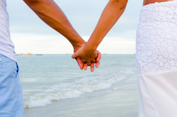 Couple holding hands having the sea as a background. View of the arms in the center and part of the bodies in the sides. A wave forming a little foam.