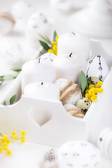 Beautiful Easter composition with white unlit candles in a white wooden box with decorated Easter eggs, olive branches and yellow mimosa flowers as a symbol of spring