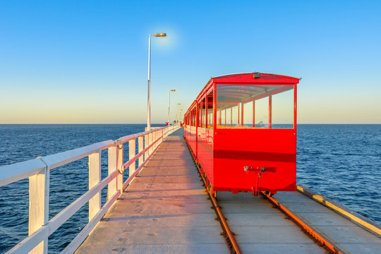 View from behind of vintage red train illuminated by sunset light, going on Jetty Busselton tracks in Busselton town, WA. Busselton Jetty is the longest wooden pier in the world and iconic place.