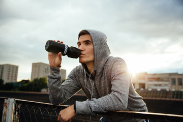 close-up portrait of handsome sportsman drinking a water after work out, listening to music in headphones, looking to the side, in hood, outdoors