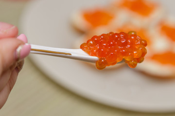 On a white spoon there is red caviar in the background of a plate with sandwiches which is not in focus. Close-up, side view