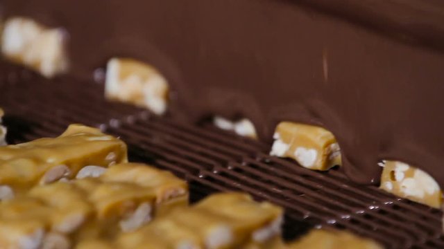Bars of nougat with nuts are poured with chocolate. Manufacture of chocolate bars with nougat and peanuts. Chocolate bars move along conveyor belt at confectionery factory for the production of sweets