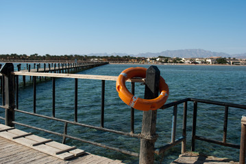 Orange lifebuoy on barrier pillar. Save lifebuoy and blue water. Safety equipment on dock for emergency. Lifebuoy and pier. Security on water.