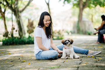 Portrait of young Pan Asian woman with her puppy dog in a park. They are sitting in the park outdoors in the sun during the day and playing with one another. 
