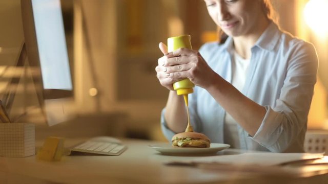 Cinemagraph of young woman squeezing out extra mustard on hamburger while sitting at work table