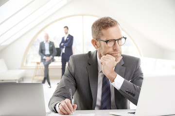Businessman using laptop at the office. Portrait of senior financial male director wearing suit and tie sitting at desk and working. Uncrecognizable business people standing at the background. 