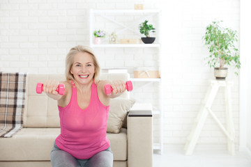 Deep squat. Front view of middle aged woman in sportswear doing squat and holding dumbbells while standing in front of window at home.
