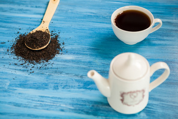 Cup with tea next to a wooden spoon with tea leaves on vintage blue background