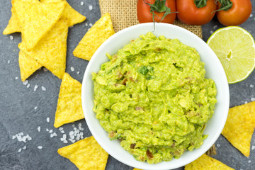 Avocado dip guacamole with tortilla chips in a white bowl on a black stone table. Traditional Mexican appetizer.